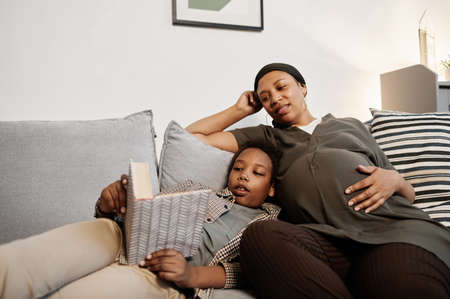 Portrait of pregnant African-American woman reading books with teenage son while relaxing at home togetherの写真素材