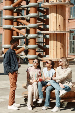 Vertical full length portrait of diverse group of young people chatting while rrelxing in urban city...