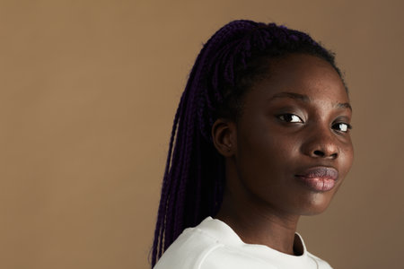 Minimal close up portrait of young African-American woman looking at camera against beige background, copy spaceの写真素材