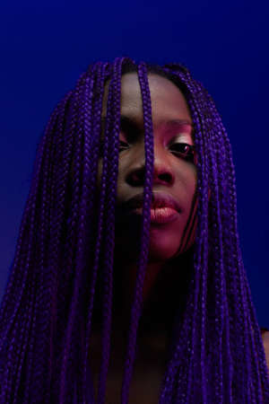 Vertical close up portrait of young woman with afro braids looking at camera against purpleの写真素材