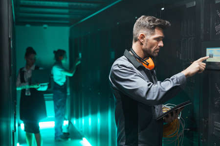 Side view portrait of male network engineer using control panel while operating server in data...