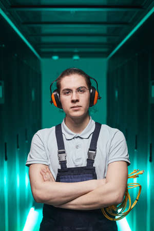 Vertical waist up portrait of young network technician looking at camera while standing with arms crossed in server roomの写真素材