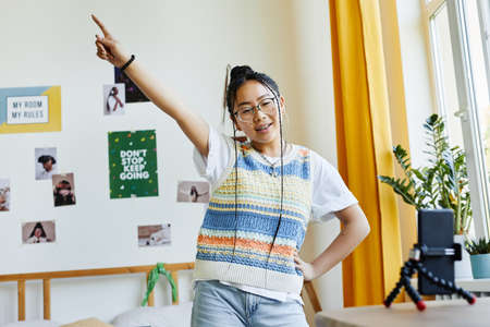 Waist up portrait of trendy teenage girl dancing to camera while filming videos in cozy room...