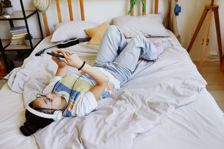 High angle view at trendy teenage girl laying on bed and using smartphone in cozy room interior, copy spaceの写真素材