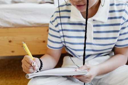 Close up of teenage girl writing in notebook while sitting on floor at home and doing homework, copy...