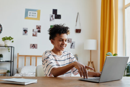 Side view portrait of mixed-race teenage boy using laptop and smiling in bright room interior, copy spaceの写真素材