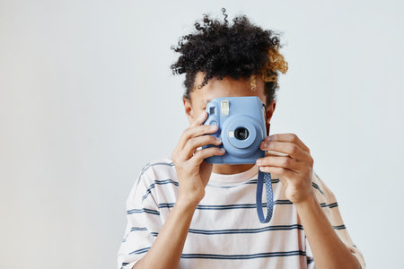 Minimal portrait of mixed-race teenage boy holding instant camera against white background, copy...