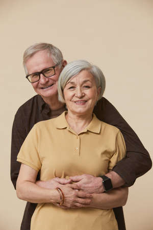 Minimal waist up portrait of senior couple embracing and looking at camera while standing against...