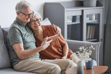 Portrait of modern senior couple using tablet at home and smiling happily while sitting on couch in minimal interior, copy spaceの写真素材
