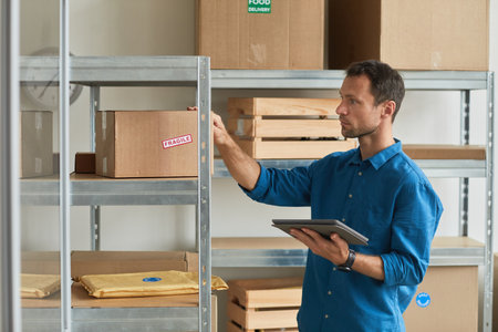 Side view portrait of young man using digital tablet in warehouse while doing inventory and managing...