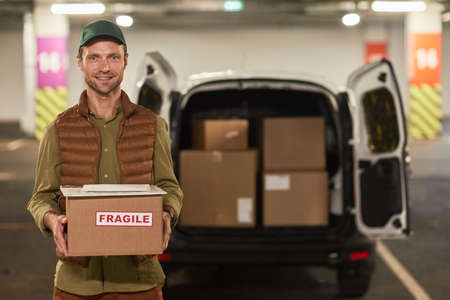 Waist up portrait of male delivery worker holding boxes and smiling at camera while standing by van...