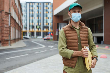 Waist up portrait of delivery man wearing mask and looking at camera while posing outdoors, copy...