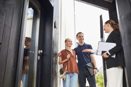 Waist up portrait of female real estate agent meeting young couple standing in doorway while buying...