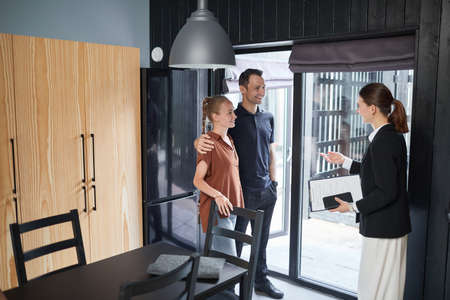 Full length side view portrait of young couple talking to real estate agent while buying new house,...