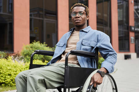 Pensive young African-American man with paralyzed legs using wheelchair while being on stroll