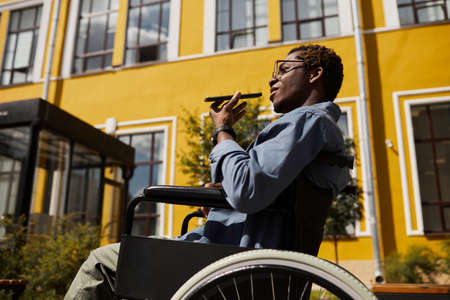 Confident disabled young black enreprenuer in eyeglasses sitting in wheelchair against yellow building and recording audio messageの写真素材