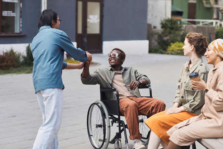 Group of young multi-ethnic friends including disabled man in wheelchair gathering together outdoors, men making handshakeの写真素材