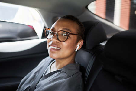 Portrait of African businesswoman in eyeglasses sitting on back seat of the car and smiling at...