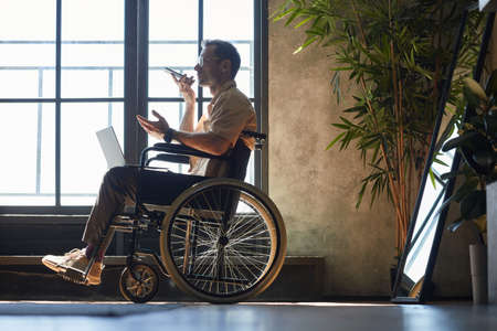 Full length portrait of modern adult man in wheelchair using smartphone while working at home lit by...