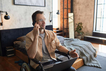 Portrait of modern adult man with disability speaking by smartphone while working from home in...