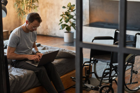 Portrait of modern disabled man using laptop while sitting on bed, wheelchair in foreground, copy...