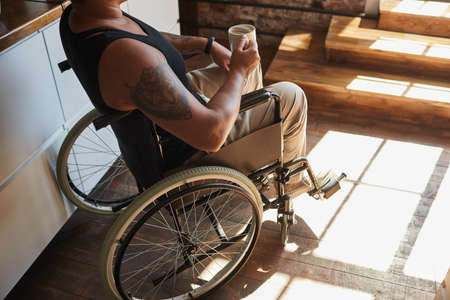 Cropped portrait of contemporary tattooed woman in wheelchair enjoying coffee at home lit by sunlight, copy spaceの写真素材