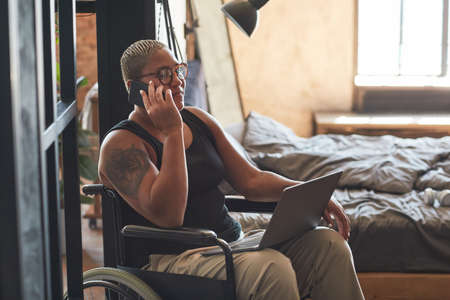Portrait of contemporary tattooed woman with disability speaking by smartphone while working from...