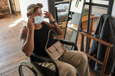 Portrait of African-American woman in wheelchair putting on mask ready to go out, copy space