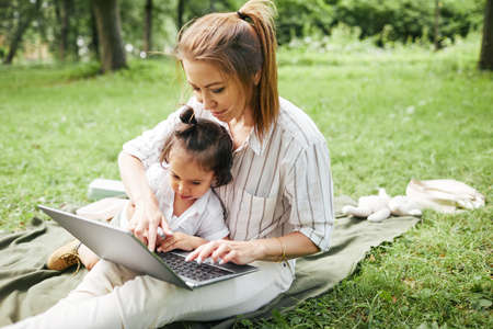 Portrait of young mother using laptop outdoors while playing with cute daughter in park, copy space