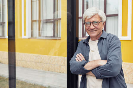 Waist up portrait of smiling mature man posing outdoors in city while standing with arms crossed and...
