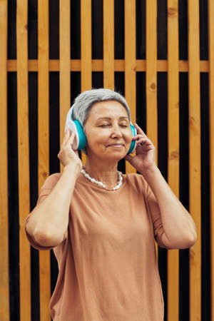 Vertical portrait of modern mature woman listening to music outdoors with eyes closed while wearing big headphones against graphic backgroundの写真素材