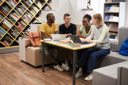Full length view at diverse group of young people studying together at table in college library,...