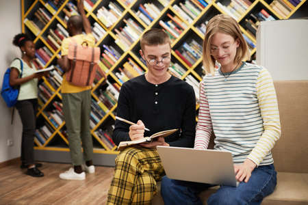 Portrait of two cheerful students using laptop together while working in college library, copy spaceの写真素材