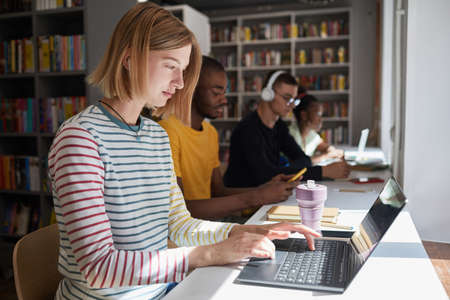 Side view portrait of young woman studying with group of students in school library, copy space
