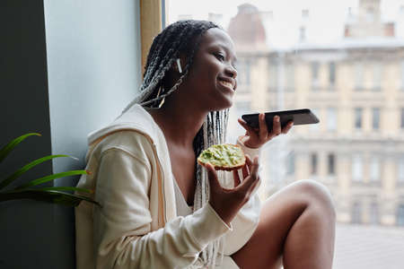 Side view portrait of young African-American woman recording voice messages while sitting by window...