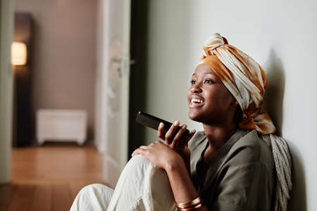Side view portrait of young African-American woman recording voice message while sitting on floor at...