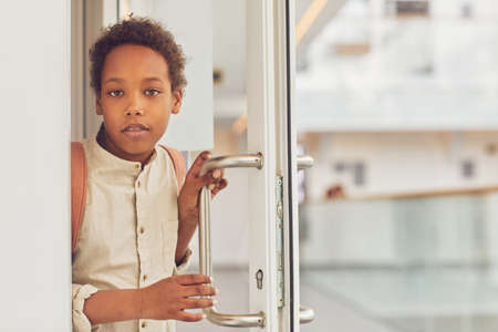 Waist up portrait of young African-American schoolboy looking at camera while opening door to classroom, copy spaceの写真素材