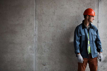 Dramatic portrait of modern construction worker looking away while standing against concrete wall,...