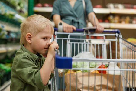Tired little blond-haired boy in green shirt leaning on shopping cart while his mother buying products in supermarketの写真素材