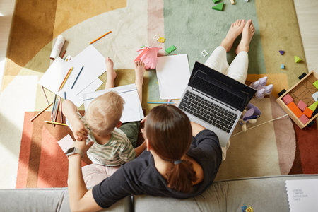 Directly above view of multitasking mother sitting on floor while working and drawing with son at same timeの写真素材
