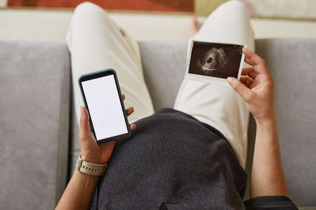 Above view of unrecognizable pregnant woman using smartphone while sending photo of ultrasound image...