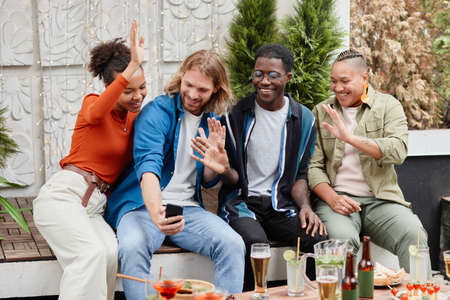 Diverse group of young people waving at camera via video chat while having fun during outdoor party...