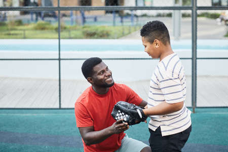 Happy handsome African-American father with beard chatting with teenage son in baseball glove while...