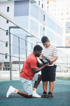 Serious black baseball coach being down on one knee and adjusting glove on boys hand at sports groundの写真素材