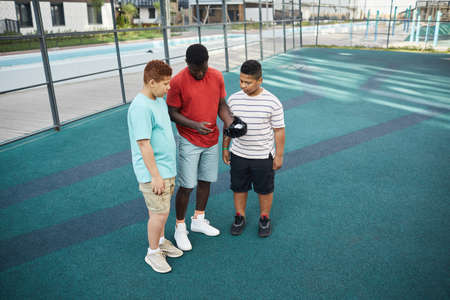 Black trainer showing hand position while giving advices about baseball game to teenage boys on sports groundの写真素材