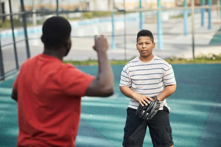 Rear view of black father throwing ball to son while playing baseball with him at playground