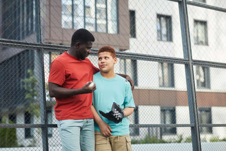 Careful African-American father in orange tshirt embracing shoulder of son and holding ball while talking about baseball to himの写真素材