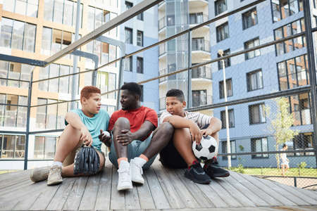 Smiling young Black man sitting on wooden podium aganinst block flat building and telling about sport to young brothersの写真素材