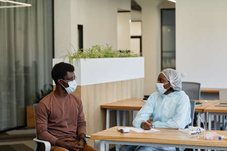Medical nurse in protective suit and medical mask talking to patient before he can get vaccine against COVID-19の写真素材