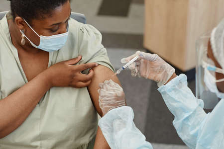 Serious African-American woman in medical mask looking at nurse getting shot of vaccine against covid-19の写真素材